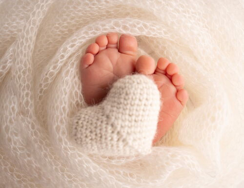 Nahaufnahme von Babyfüßen, die in einem weißen, weichen Tuch eingewickelt sind, mit einem gestrickten weißen Herz zwischen den Füßen
